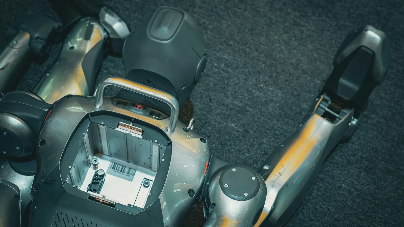 Torso de un robot humanoide tumbado boca abajo sobre moqueta oscura, con una compuerta abierta que deja ver los componentes internos