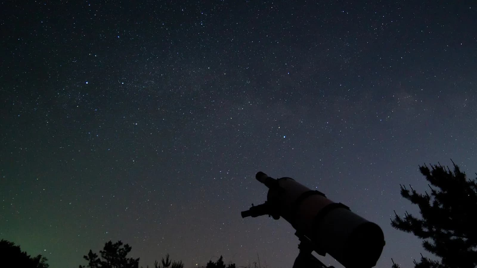 Silueta de un telescopio apuntando a un cielo nocturno lleno de estrellas, con un tenue resplandor verdoso en el horizonte.