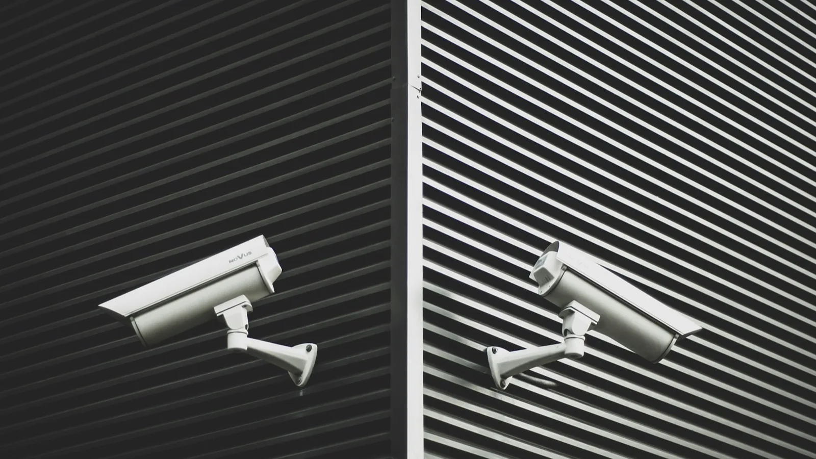 Dos cámaras de videovigilancia blancas montadas en una fachada metálica de listones oscuros, una sobre fondo negro y otra sobre fondo claro