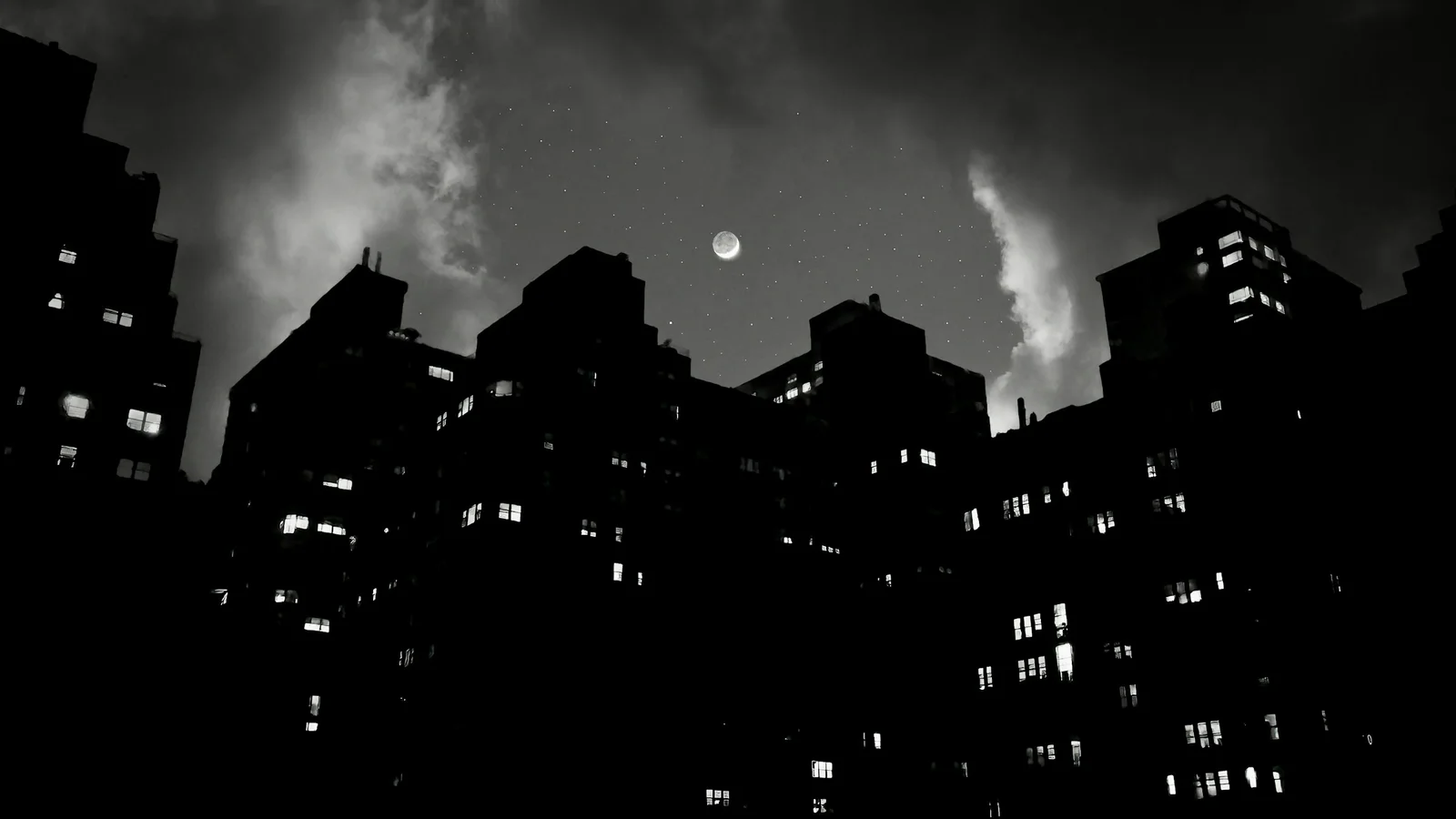 Siluetas de edificios bajo un cielo nocturno nublado, con la luna entre las nubes.