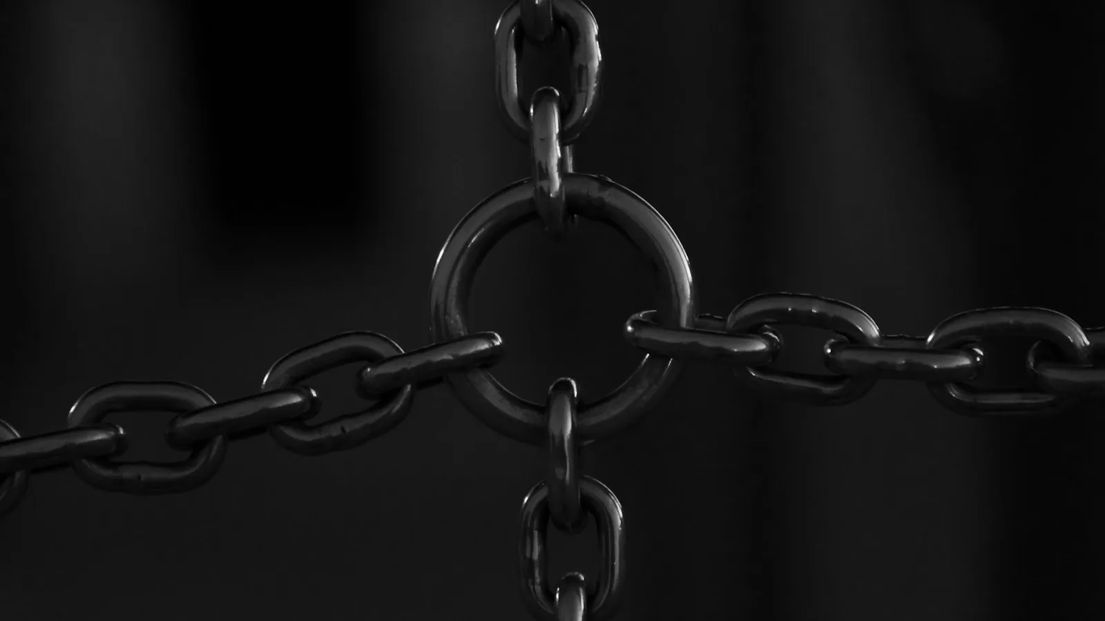 Primer plano en blanco y negro de varios eslabones de una cadena metálica convergiendo en una argolla central, sobre fondo oscuro