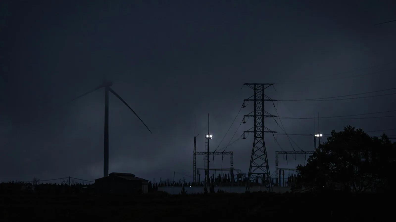 Silueta nocturna de una torre de alta tensión y un aerogenerador junto a una pequeña subestación eléctrica iluminada, bajo un cielo oscuro y nublado