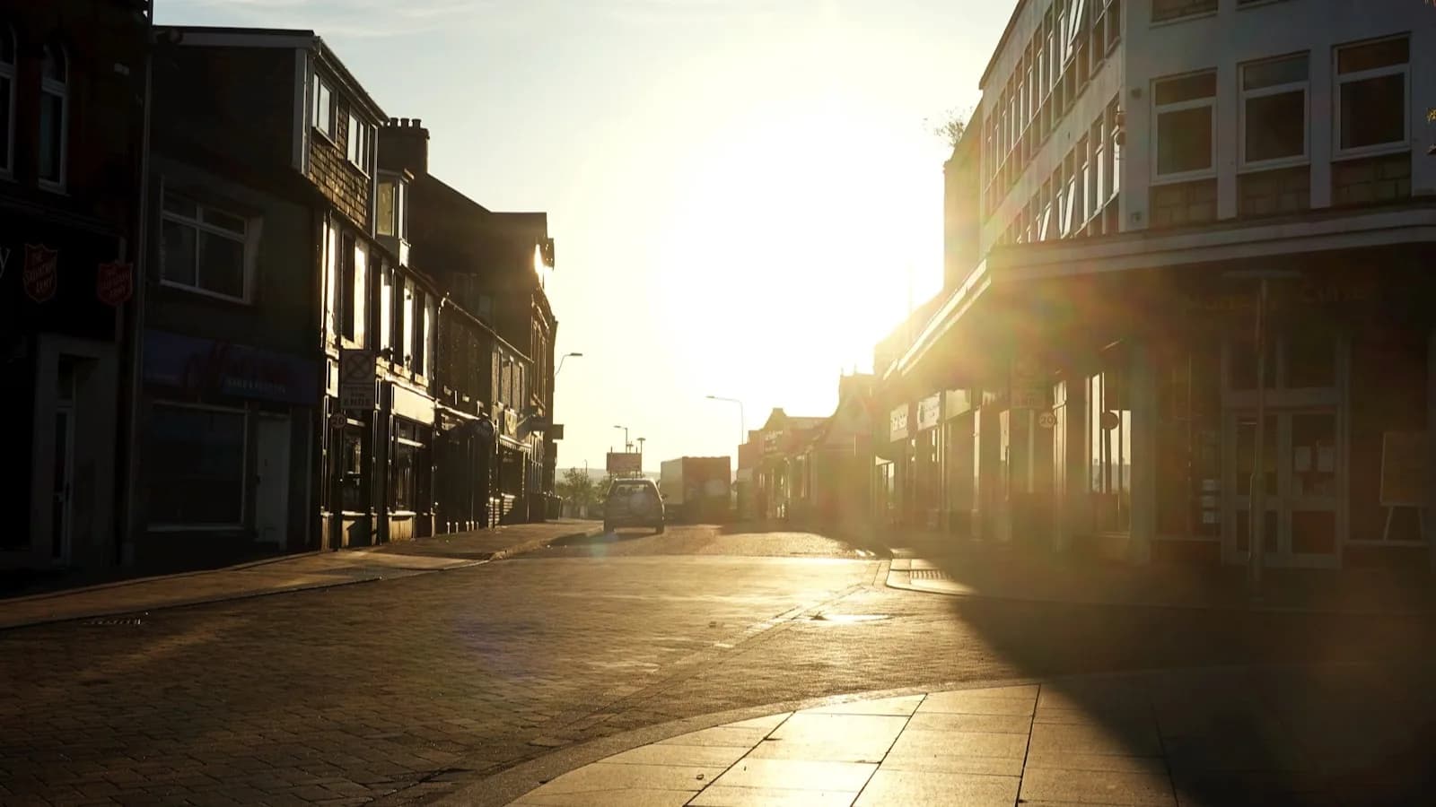 Una calle comercial vacía al atardecer con el sol bajo proyectando largas sombras entre los edificios