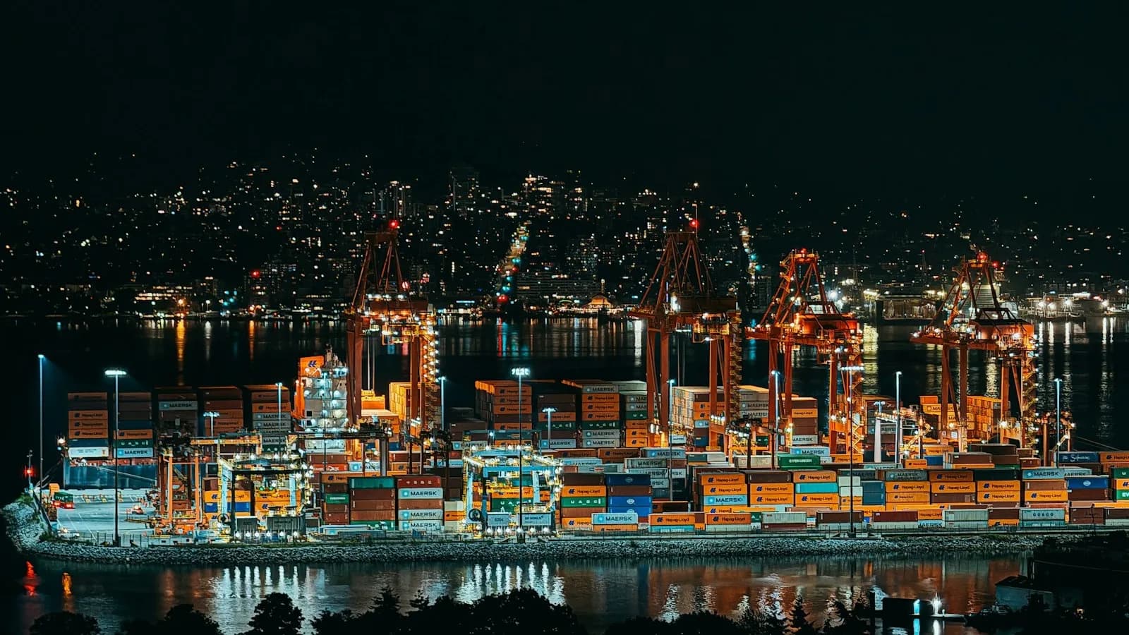 Terminal portuaria de noche con grúas y miles de contenedores apilados bajo focos, con una ciudad iluminada al fondo