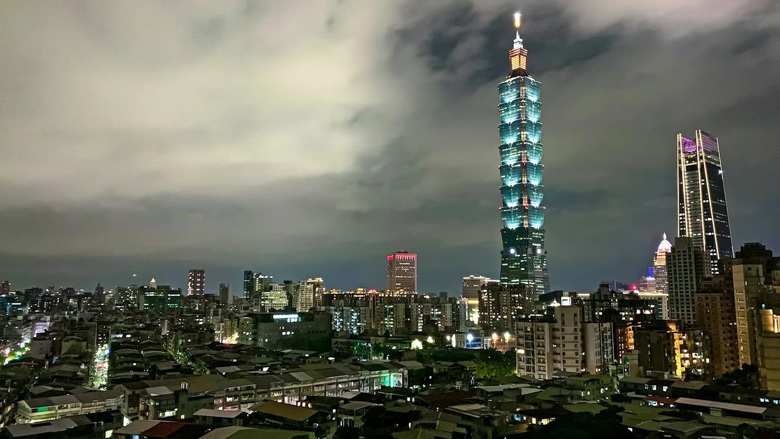 Vista nocturna del rascacielos Taipei 101 iluminado en azul, destacando sobre el resto de la ciudad bajo un cielo nublado