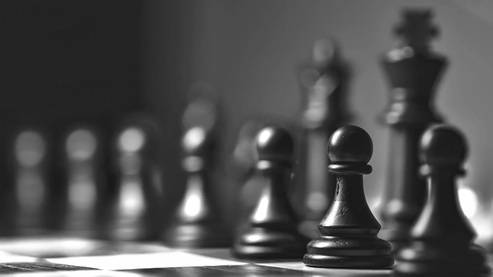 Piezas de ajedrez negras en blanco y negro, con los peones en primer plano y el rey desenfocado al fondo