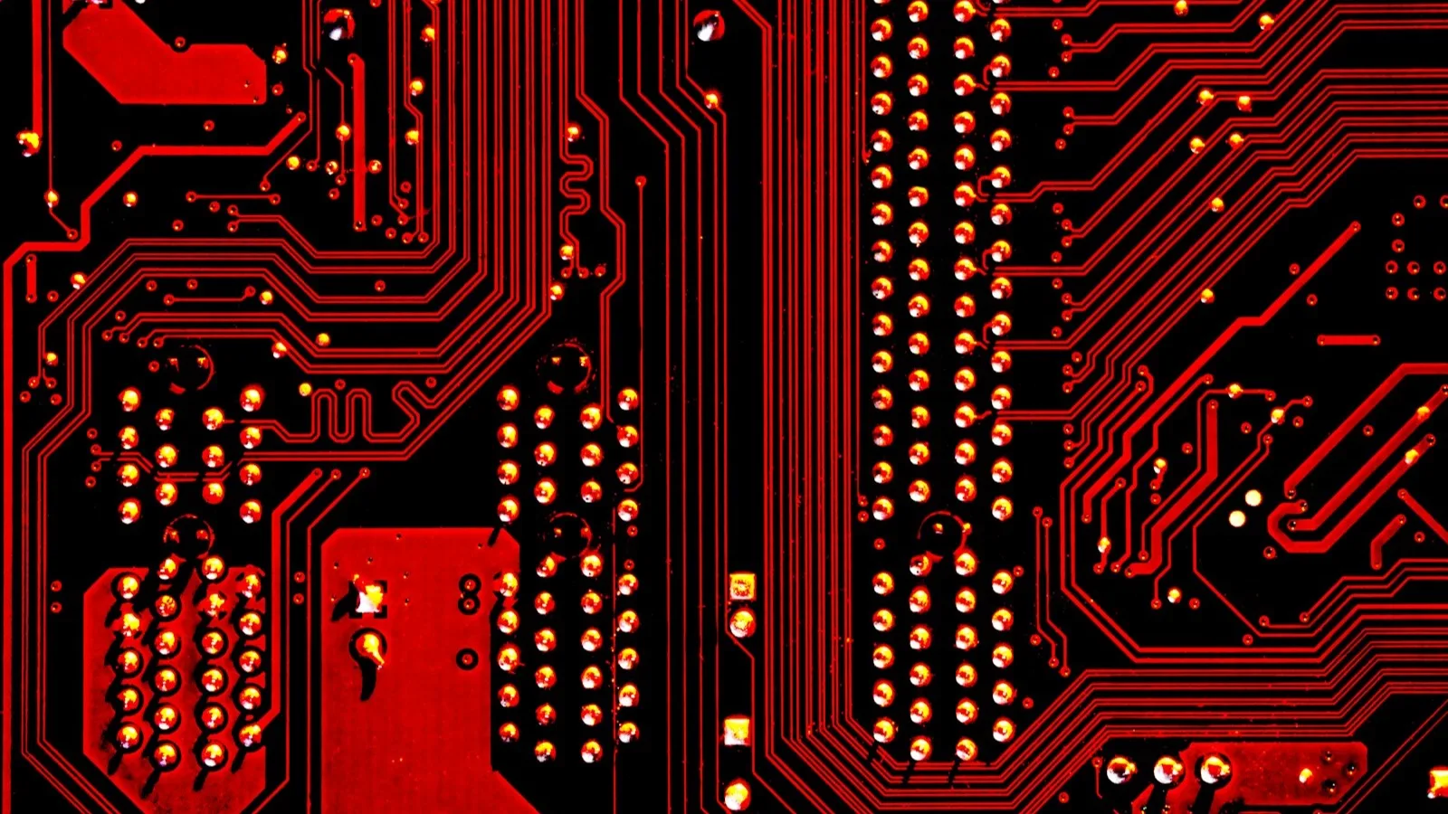 Primer plano de una placa de circuito impreso en tonos rojo y negro, con pistas metálicas y componentes soldados